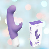 A VeDO Joy Vibe G-Spot Rabbit Vibrator, featuring dual stimulation points with a G-spot rabbit and clitoris tickler in a vibrant purple color, is displayed next to its packaging box. The box showcases an identical product image and stands out with its white design accented by purple highlights and text. The background is adorned with a blurred, colorful bokeh effect.